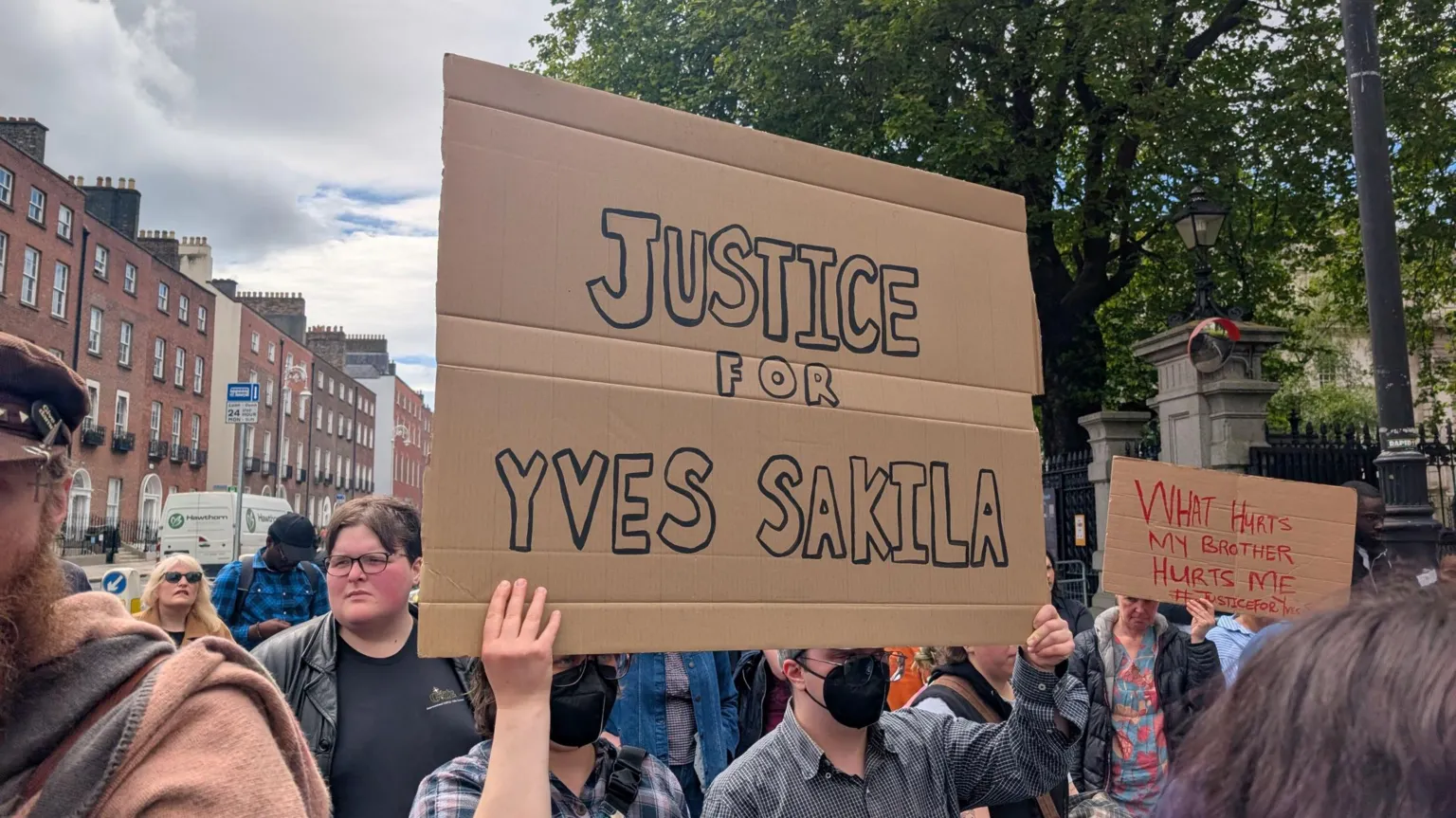 PA Media Two people wearing black face masks, hold up a cardboard sign which reads 'Justice for Yves Sakila' in black writing. They stand among a group of people in the shade of large green trees.
