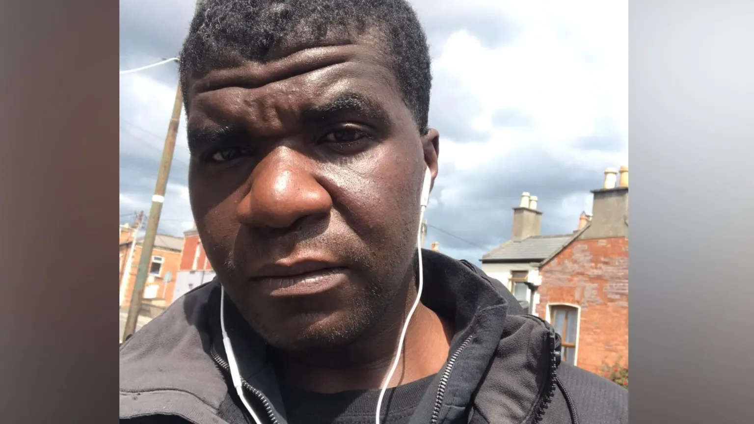 Social media A man looks into the camera with a neutral expression. His name is Yves Sakila, originally from the DR Congo. He is outside on a street, in the background terraced houses can be seen along with a cloudy sky. He is wearing a black coat and has white wired earphones in his ears. 