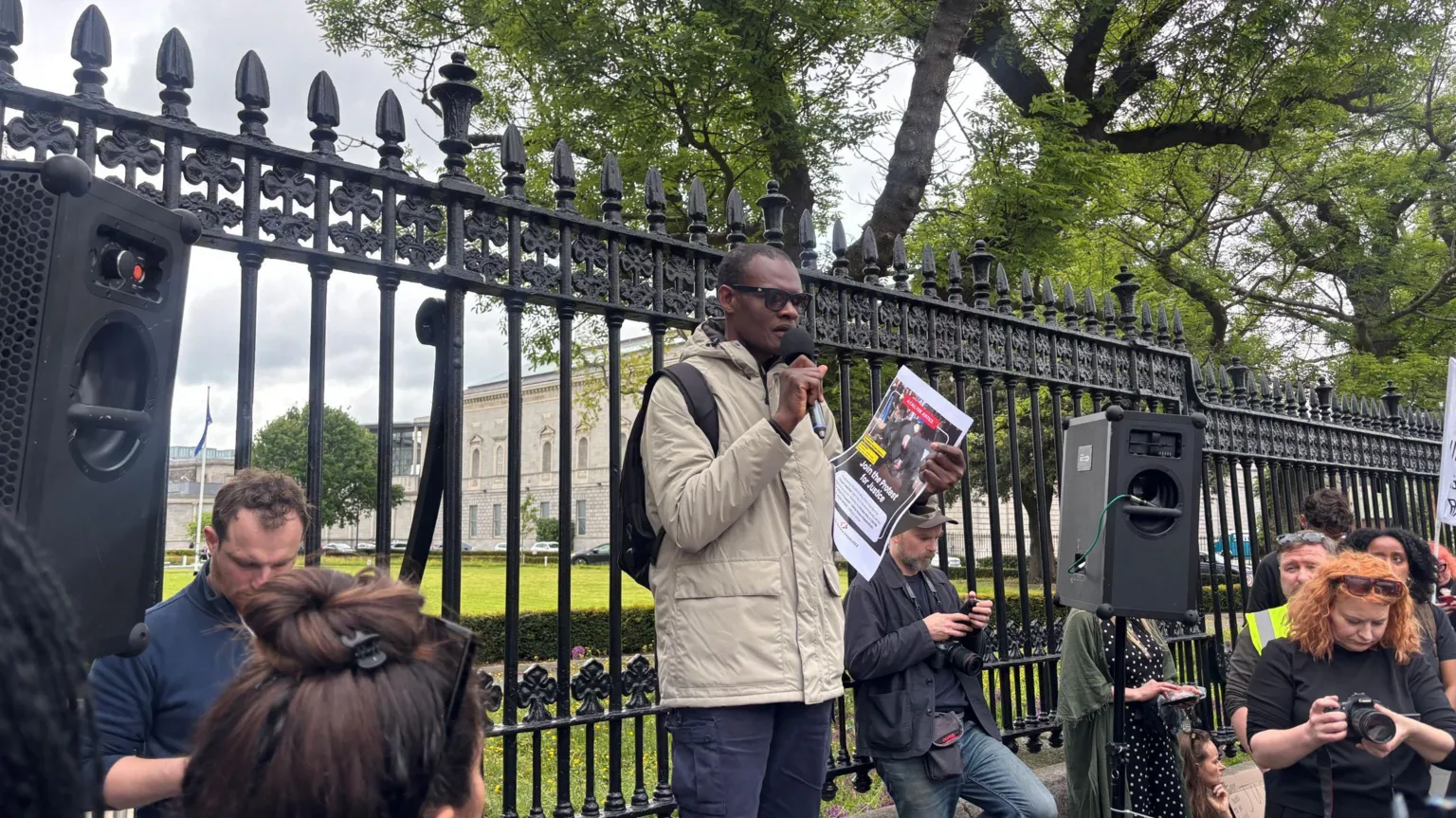 A man with short black hair and sunglasses wears a cream jacket and navy jeans. He holds a microphone in his right hand. There is a black speaker to his left. He stands in front of the black railings that sit in front of the Irish parliament. 