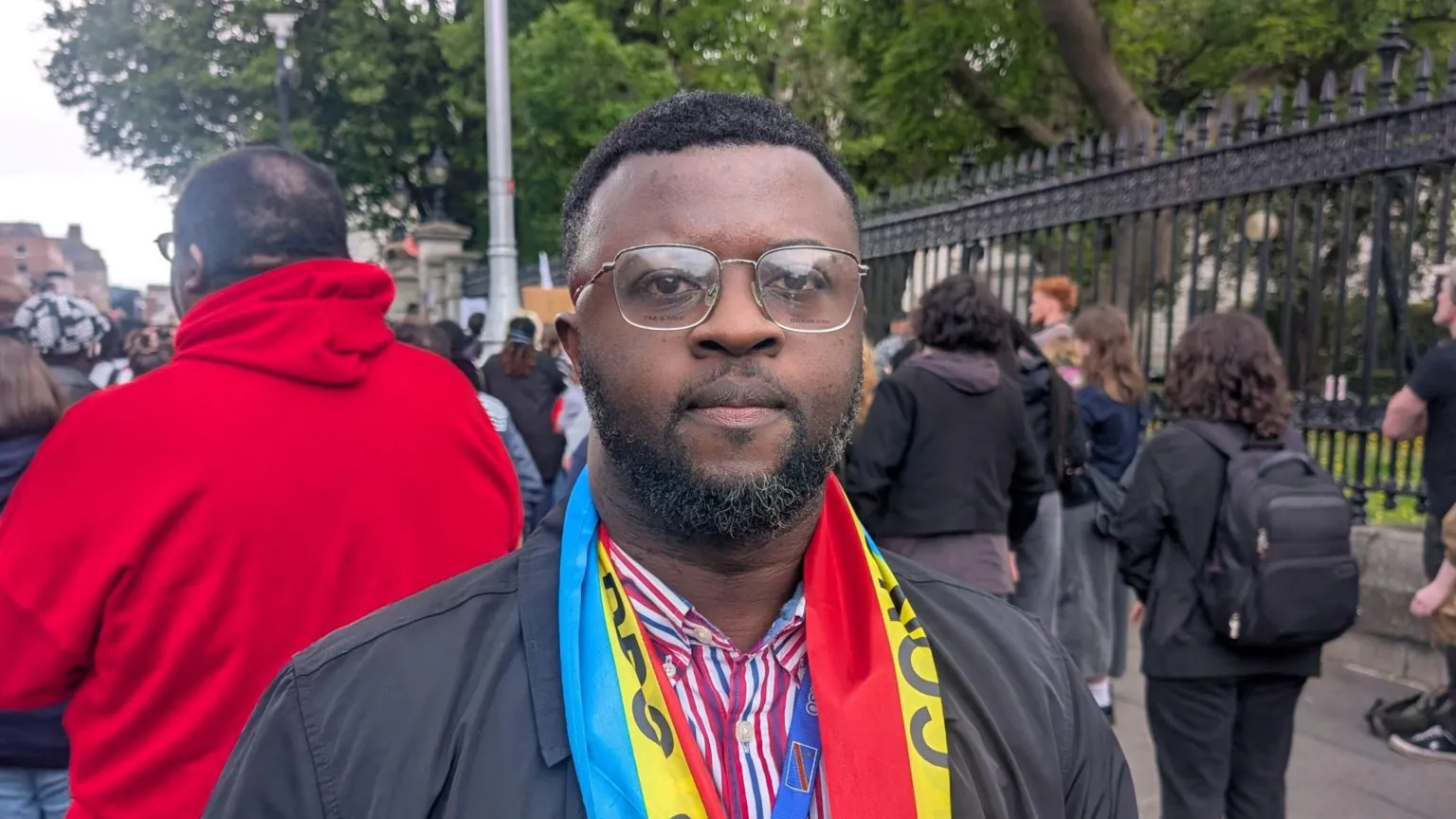 PA Media A man stands in a black coat and blue and white striped shirt. He has short black hair and a short black beard. He wears a yellow, blue and red scarf that reads 'Congo' on it. 